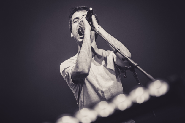 Ein Mann in schwarzem T-Shirt und Jeans singt leidenschaftlich in ein Mikrofon auf einer hell erleuchteten Bühne, die Augen geschlossen, mit dramatischer Beleuchtung.