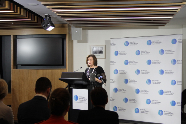 Eine Frau steht auf einer Bühne und spricht in ein Mikrofon, vor ihr sitzen Menschen auf Stühlen, an der Wand hinter ihr eine Fahne, ein Fernseher an der hölzernen Wand und Lampen an der Decke.