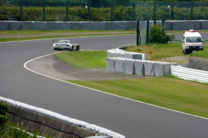 Ein Rennwagen fährt eine Rennstrecke entlang mit einem Polizeiwagen im Hintergrund, umgeben von Gras, Pflanzen, Bäumen und einem Fahrzeug auf der rechten Seite und Text unten.