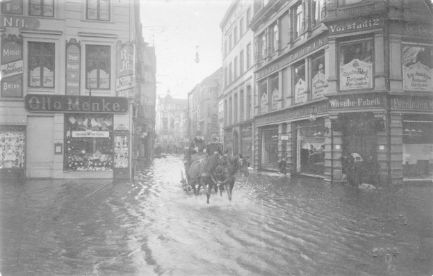 Ein von Pferden gezogenes Fuhrwerk fährt durch eine überflutete Straße in Berlin, Deutschland, mit Gebäuden auf beiden Seiten und einem schwarz-weißen Himmel im Hintergrund.