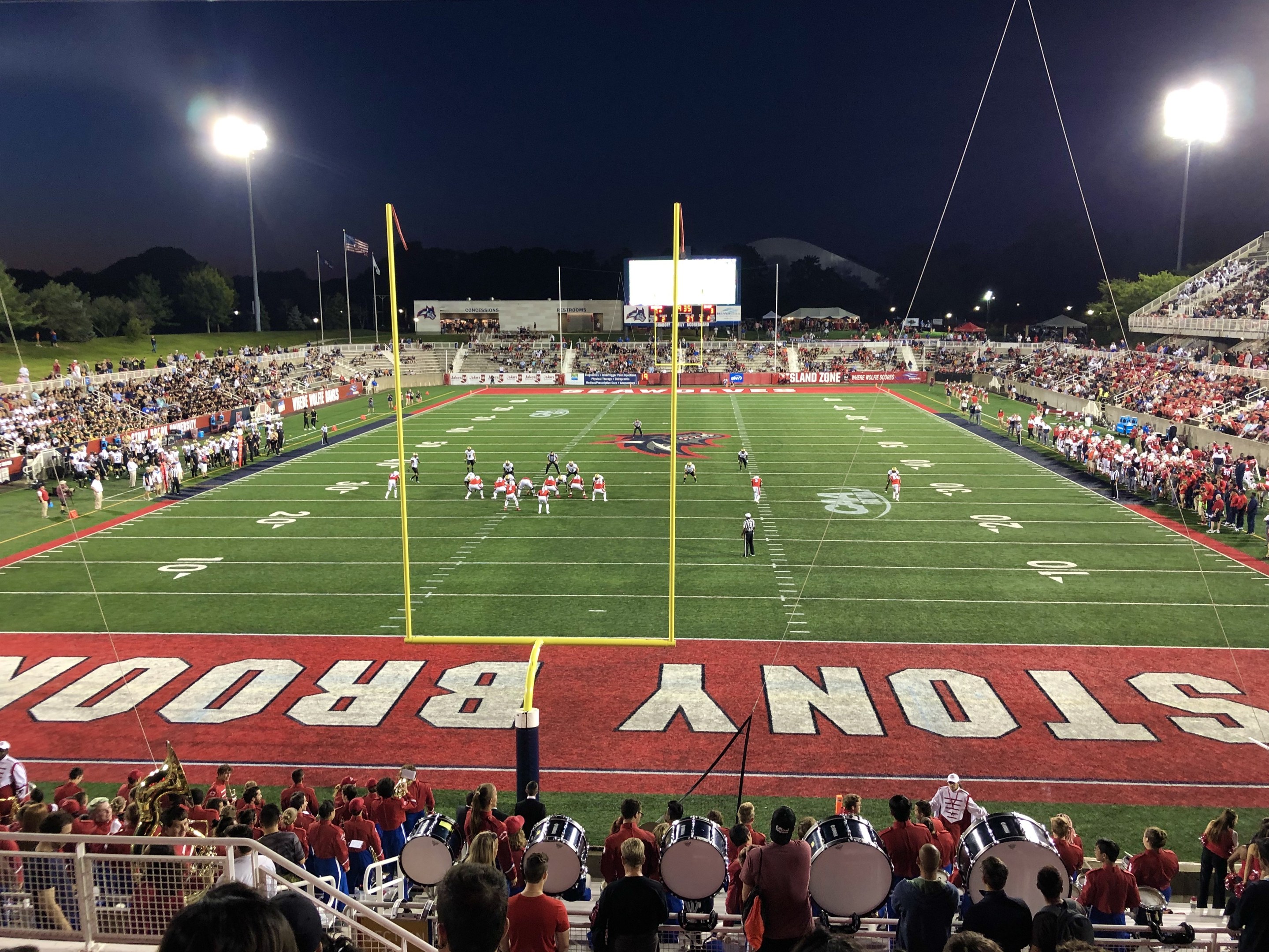 Ein nächtliches Fußballspiel in einem Stadion mit Spielern auf dem Feld, Zuschauern auf Stühlen und Musikern, umgeben von einem Metallzaun, mit Laternen, Fahnen, einem Display, Bäumen, Hügeln und einem sternenklaren Himmel im Hintergrund.