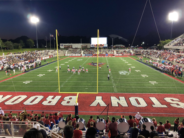 Ein nächtliches Fußballspiel in einem Stadion mit Spielern auf dem Feld, Zuschauern auf Stühlen und Musikern, umgeben von einem Metallzaun, mit Laternen, Fahnen, einem Display, Bäumen, Hügeln und einem sternenklaren Himmel im Hintergrund.