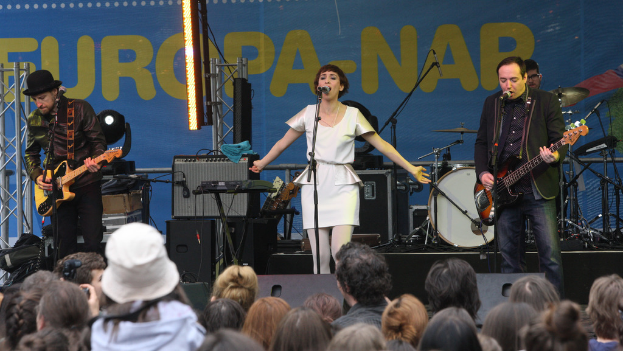 Eine Bühne mit vier Musikern: eine singende Frau in weißem Kleid in der Mitte, zwei Gitarristen links und rechts und Publikum und Lautsprecher im Hintergrund.