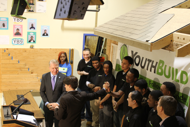 Eine Gruppe von Menschen, die um ein Rednerpult herumstehen und vor einer Menge, mit einem Tisch mit Mikros und Papieren links daneben, Plakaten an der Wand im Hintergrund und einem Objekt auf der rechten Seite, sich die Hände schütteln, was darauf hinweist, dass sie Teil einer Jugendgruppe sind.