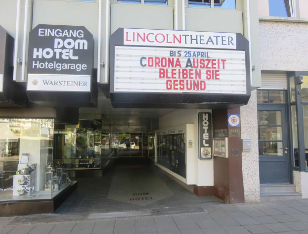 Außenansicht des Lincoln Theaters in Berlin, Deutschland, mit Glasfenstern und -türen sowie einer Texttafel und einem belebten Stadtbild im Inneren.
