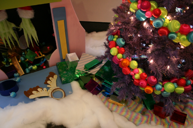 Drinnen steht ein geschmückter Weihnachtsbaum mit Lichterkette und Schmuck rechts, Geschenkboxen und dekorative Gegenstände in der Mitte, eine Hütte mit Dekoration oben links und Watte und Wolle unten.