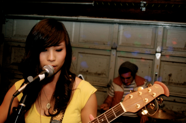 Eine Frau spielt Gitarre und singt auf der Bühne mit einem Mikrofon links von ihr, während ein sitzender Mann im Hintergrund an einer Wand zu sehen ist.