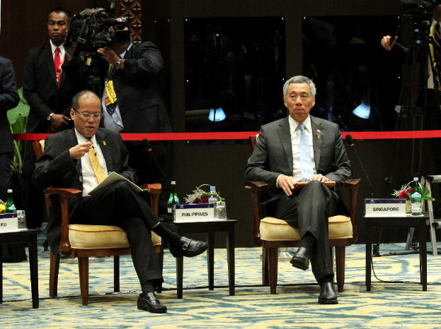 Zwei Personen, eine liest ein Buch und spricht, sitzen an einem Tisch mit einer Wasserflasche und Namensschildern, während Kameramann im Hintergrund filmt.