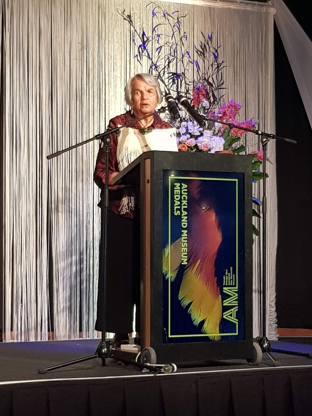 Eine Frau steht an einem Podium mit Mikrofon, Blumen und einer Texttafel vor einem Vorhang und einer Wand bei der jährlichen Verleihung des Auckland Museum of Art Awards.