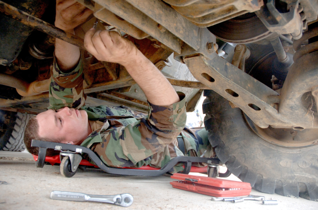 Ein Soldat liegt unter einem Fahrzeug auf dem Boden und hält einen Schraubenschlüssel in der Hand, umgeben von verschiedenen Werkzeugen.