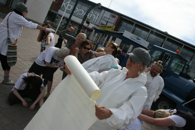 Eine Gruppe von Menschen, einige in Kostümen und Mützen, versammelt sich in der Mitte, während einige zu kämpfen scheinen, während eine Person ein Papier hält; im Hintergrund sind Gebäude, Pflanzen, Pfosten und Schilder mit Text, ein Fahrzeug auf der rechten Seite, ein Weg am Boden und der Himmel oben zu sehen.