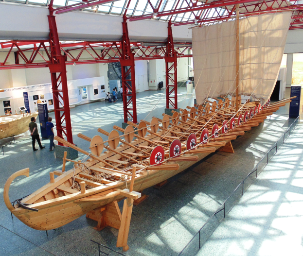 Ein großes hölzernes Wikingerschiff in einem Museum ausgestellt, umgeben von Besuchern, Metallstangen, einem Zaun, einer Fahne und Deckenleuchten.