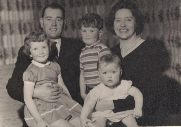 Eine Familie mit fünf Mitgliedern, einschließlich eines Mannes in Anzug und Krawatte auf der linken Seite, einer Frau auf der rechten Seite und drei strahlenden Kindern, die gemeinsam posieren.