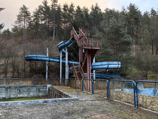 Ein verlassener Wasserpark mit einer blauen Wasserrutsche in der Mitte, umgeben von Geländern, Treppen und einer Wand, mit Bäumen und einem klaren blauen Himmel im Hintergrund und Text am unteren Bildrand.