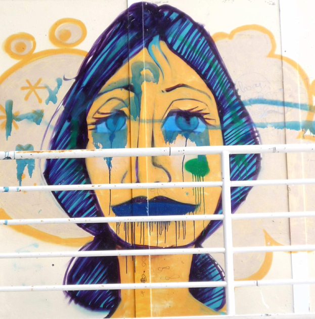 Ein Wandgemälde eines traurigen Frauenkopfs an einem Gebäude in Berlin, mit einem Gitter im Vordergrund, gemalt in gedämpften, melancholischen Farben.