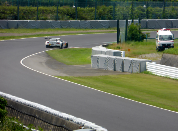 Ein Rennwagen fährt eine Rennstrecke entlang mit einem Polizeiwagen im Hintergrund, umgeben von Gras, Pflanzen, Bäumen und einem Fahrzeug auf der rechten Seite und Text unten.