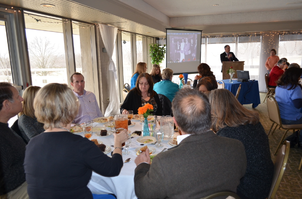 Eine Gruppe von Menschen sitzt um einen Tisch, der mit verschiedenen Speisen gefüllt ist, eine stehende Person spricht, ein Projektor zeigt Inhalte, eine Pflanze neben dem Projektor und ein Blick aus dem Fenster mit Vorhängen, Himmel und Bäumen im Hintergrund.
