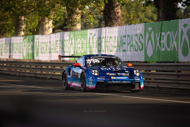 Ein blauer Porsche 911 GTS auf einer Rennstrecke, umgeben von einem Zaun und Bannern mit Text, mit B├Ąumen und einem klaren blauen Himmel im Hintergrund.