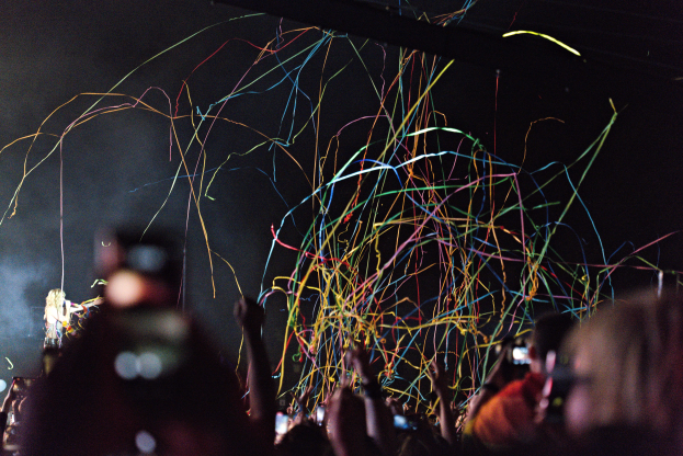 Eine Menge bei einem Konzert mit erhobenen Händen, beleuchtet von bunten Lichtern, mit einer Person, die links ein Mikrofon hält und einer anderen, die rechts eine Kamera hält.