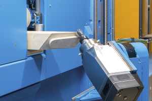 Eine CNC-Fräsmaschine schneidet ein Holzstück in einer Fabrikumgebung, umgeben von Kabeln und anderen Gegenständen, mit einer Wand im Hintergrund.