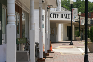 Ein Rathaus umgeben von Gebäuden, Straßenlaternen, Straßenschildern, Autos, Verkehrskegeln, Stühlen, Topfpflanzen, Bäumen und einer bewölkten Himmel.