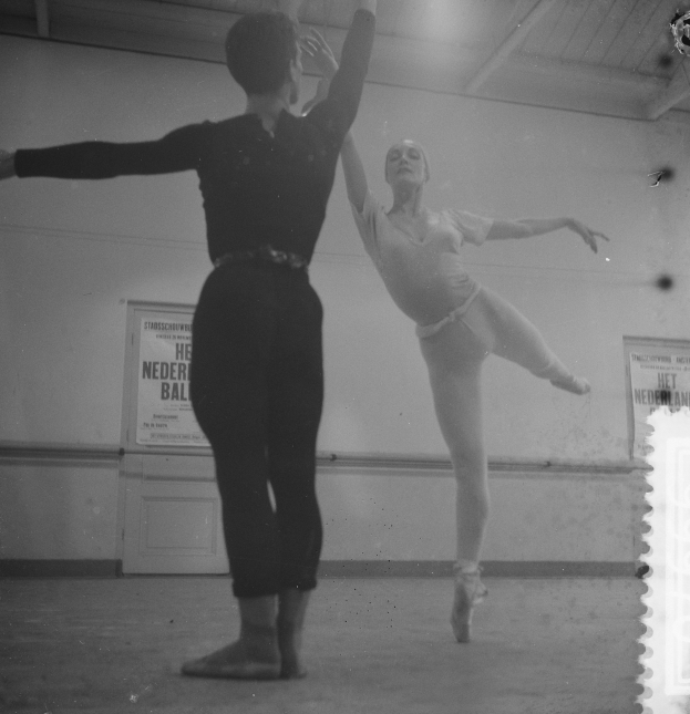 Ein Schwarz-Weiß-Foto von einem Mann und einer Frau, die in einem Studio tanzen, wobei die Frau in der Luft ist und eine Wand mit Plakaten im Hintergrund zu sehen ist, die von Deckenlampen beleuchtet wird.