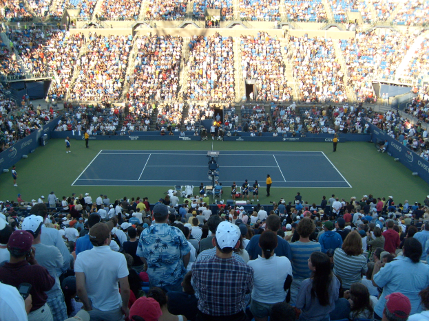 Eine große Menge Menschen in einem Stadion schaut einem Tennismatch zu, mit Spielern auf dem Platz und Zuschauern, die die Tribünen füllen und eine elektrisierende Atmosphäre schaffen.
