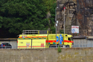 Rettungswagen auf der Seite einer Straße neben einer Klippe geparkt, mit einer Wand, einem Geländer, Pfählen, Lampen, Schildern mit Text und anderen Gegenständen im Vordergrund und Bäumen und einem Auto im Hintergrund.