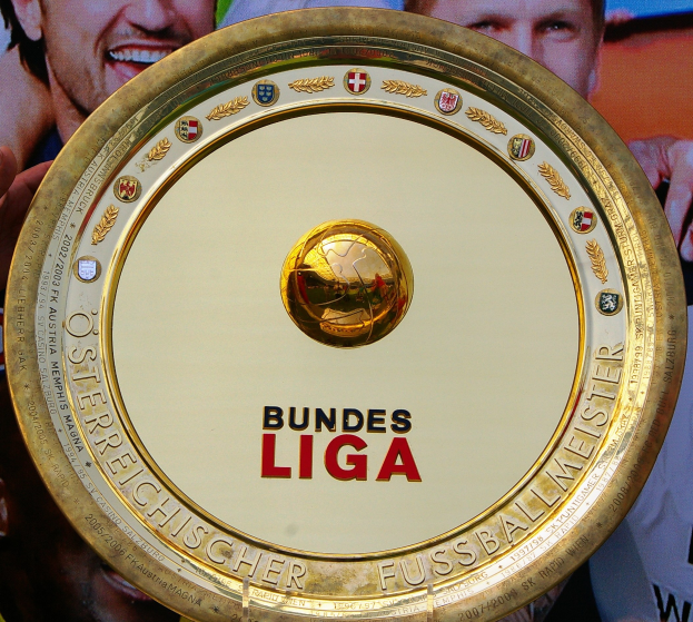 Ein Mann hält einen goldenen Schild mit der Aufschrift "Bundesliga" in die Höhe, im Hintergrund ein Banner mit drei Personen