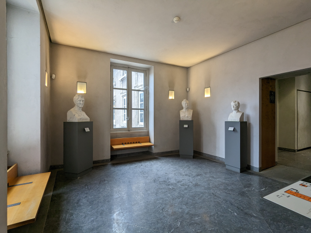 Ein Raum voller Skulpturen auf Sockeln, eine hölzerne Bank, ein Fenster, Wandlampen, eine Tür und ein Dach, das dem Museum of Fine Arts in Paris ähnelt.