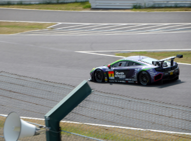 Ein Sportwagen fährt auf einer Rennstrecke mit einem unscharfen Hintergrund, umgeben von Gras und einem Zaun, mit einem Lautsprecher unten.