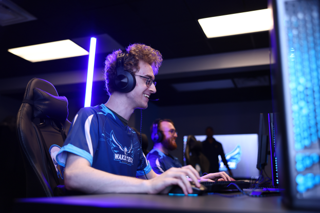 Ein Mann sitzt an einem Schreibtisch mit einem Computer, trägt einen Headset und Brille, mit einer weiteren Person und Deckenleuchten im Hintergrund, was eine gamingbezogene Umgebung im Zusammenhang mit dem Waketex Esports-Team vermuten lässt.