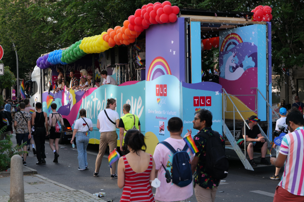 Eine Gruppe von Menschen geht eine Straße entlang neben einem Lastwagen mit bunten Luftballons, mit Schildern an der Straße und Bäumen und Gebäuden im Hintergrund, was auf eine Pride-Parade in Paris hindeutet.