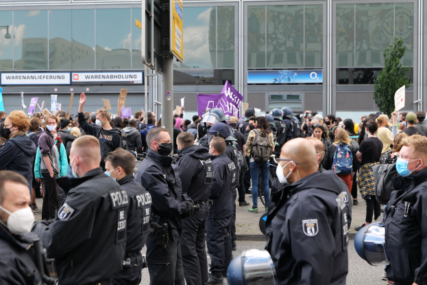 Eine große Gruppe von Menschen protestiert vor einem Gebäude, einige halten Schilder und tragen Helme, mit einem Schildständer im Vordergrund und einem Baum im Hintergrund.