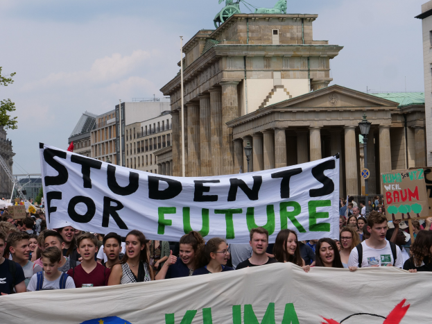 Gruppe von Studenten marschiert in Berlin mit einer bunt bemalten "Students for Future"-Schlagzeile vor einem Hintergrund aus Gebäuden, Bäumen und Himmel.