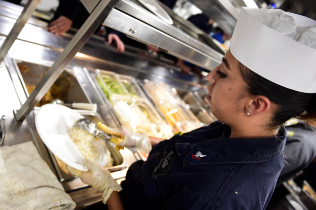 Eine Frau in Kochmütze und Handschuhen hält einen Teller mit Essen in einer Küche, mit Tabletts voller Lebensmittel auf einem drapierten Tisch und Menschen im Hintergrund.