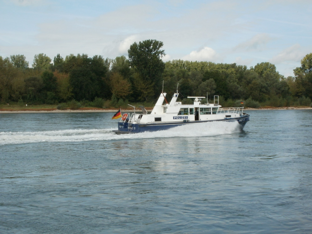 Ein Polizei-Streife-Boot mit Flaggen fährt einen Fluss mit Bäumen an den Ufern entlang unter einem klaren blauen Himmel.