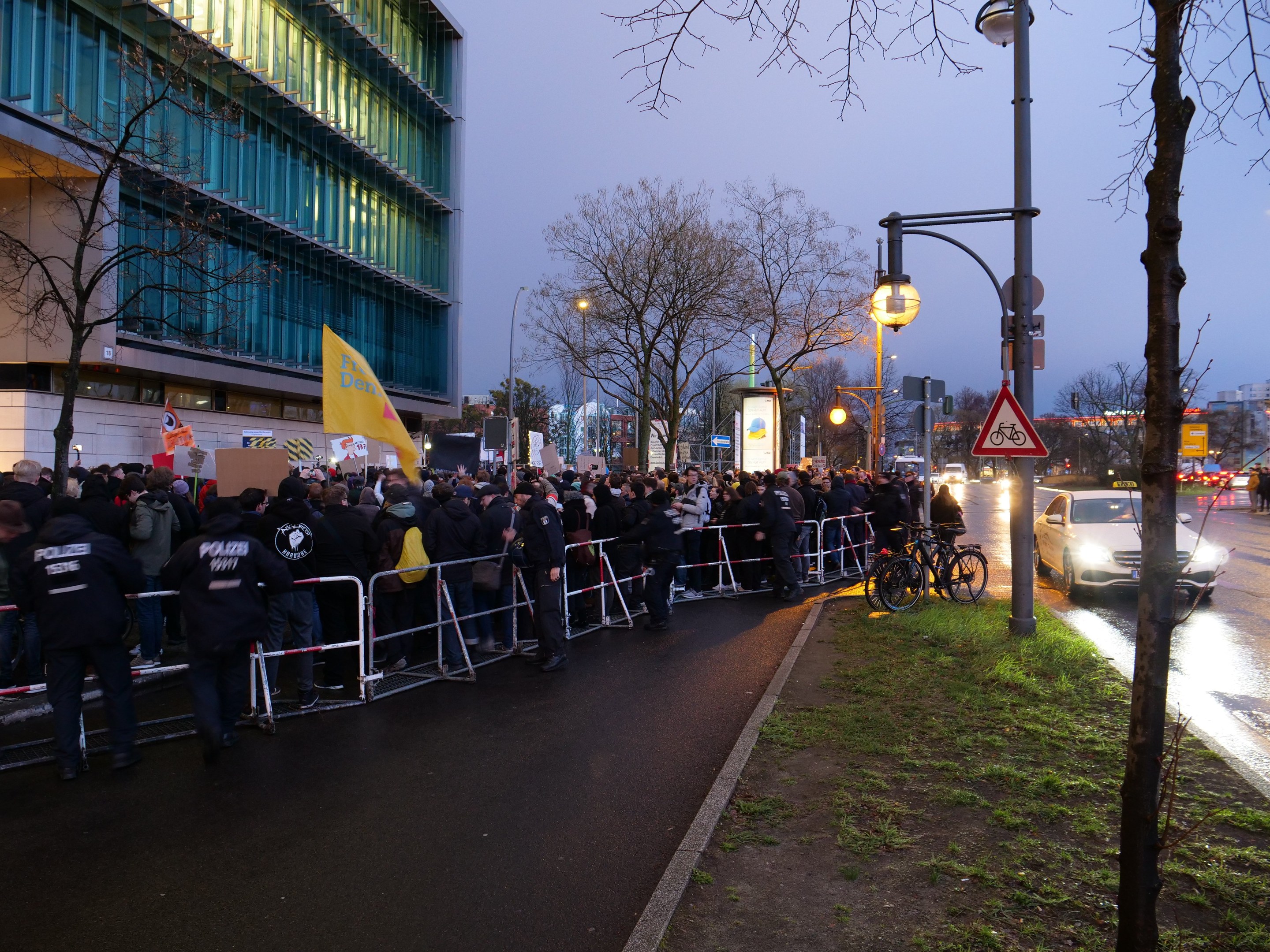 Eine große Gruppe von Menschen steht vor einem Gebäude mit Barrikaden und Schildern und hält Schilder während einer Demonstration in Berlin.