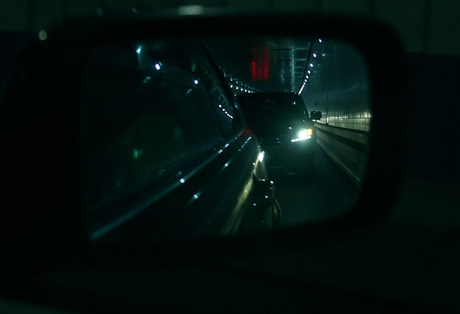 Ein Auto fährt durch einen Tunnel in der Nacht, dessen Seitenspiegel andere Fahrzeuge reflektiert, beleuchtet von Tunnel- und Autoscheinwerfern.