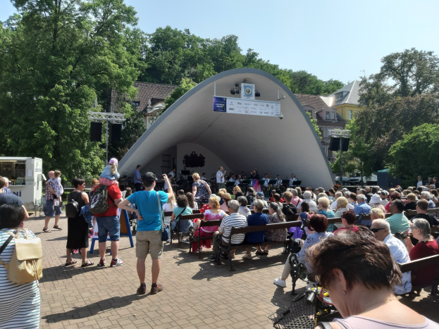 Eine Menschenmenge, die auf Bänken sitzt und vor einer Bühne steht, bei einem Outdoor-Sommerkonzert, mit einem Kameramann, einem Banner, Bäumen, Gebäuden und einem bewölkten Himmel.