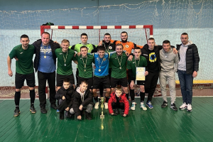 Eine Gruppe von Männern in Medaillen, einer hält einen Pokal, steht auf einem Futsal-Platz mit einem Tor und einer Wand im Hintergrund.