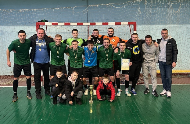 Eine Gruppe von Männern in Medaillen, einer hält einen Pokal, steht auf einem Futsal-Platz mit einem Tor und einer Wand im Hintergrund.