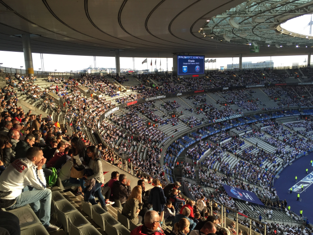 Eine große Menge Menschen in einem Stadion schaut ein Fußballspiel, mit einer Bühne rechts, Fahnen, Stangen, einem Bildschirm und der Allianz Arena in München.