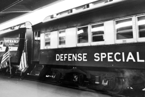 Ein Schwarz-Weiß-Foto eines stationären Zugs mit der Aufschrift 'Defense Special' und zwei Flaggen auf der linken Seite und einer Decke öberhaupt.