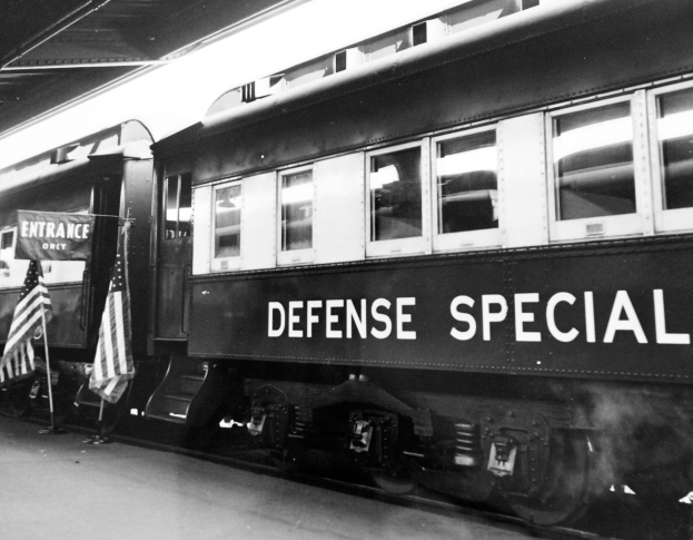 Ein Schwarz-Weiß-Foto eines stationären Zugs mit der Aufschrift 'Defense Special' und zwei Flaggen auf der linken Seite und einer Decke öberhaupt.