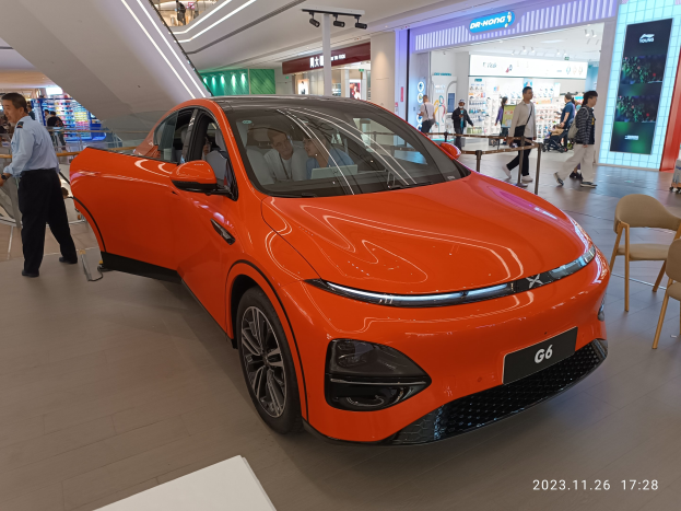 Oranger Stromwagen in einer Ausstellungshalle mit einer Gruppe von Menschen drumherum, Stühlen und einem Wagen auf der rechten Seite und Geschäften im Hintergrund.