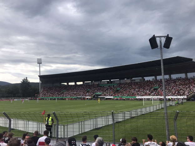 Eine große Menschenmenge schaut einem Fußballspiel in einem eingezäunten Stadion bei künstlicher Beleuchtung zu, mit Bäumen und Hügeln im Hintergrund unter einem bewölkten Himmel.