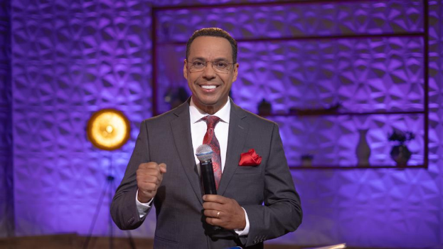 Stephen Colbert, der in einem Anzug, einer Krawatte und einer Brille auf einer Bühne steht und ein Mikrofon hält.