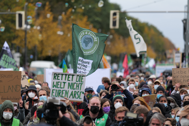 Eine große Gruppe von Menschen mit Masken und Schildern, Fahnen und Kameras nimmt an einem Klimastreik in Deutschland teil, umgeben von Bäumen, Pfählen, Verkehrszeichen und Fahrzeugen mit dem Himmel im Hintergrund.