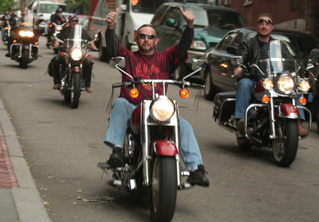 Eine Gruppe von Menschen, die auf Fahrrädern auf einer Straße fahren, wobei eine Person in einem roten Hemd ohne Hände fährt und eine andere Person in einer schwarzen Jacke lächelt. Im Hintergrund sind zwei Autos und ein Lieferwagen zu sehen.